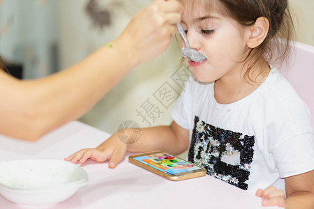 母亲在看智能电话的视频时喂小女儿吃东西图片
