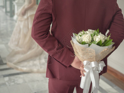 穿红色或布贡迪婚纱装的Groom在新娘背后藏着花束白色美丽的玫瑰花图片