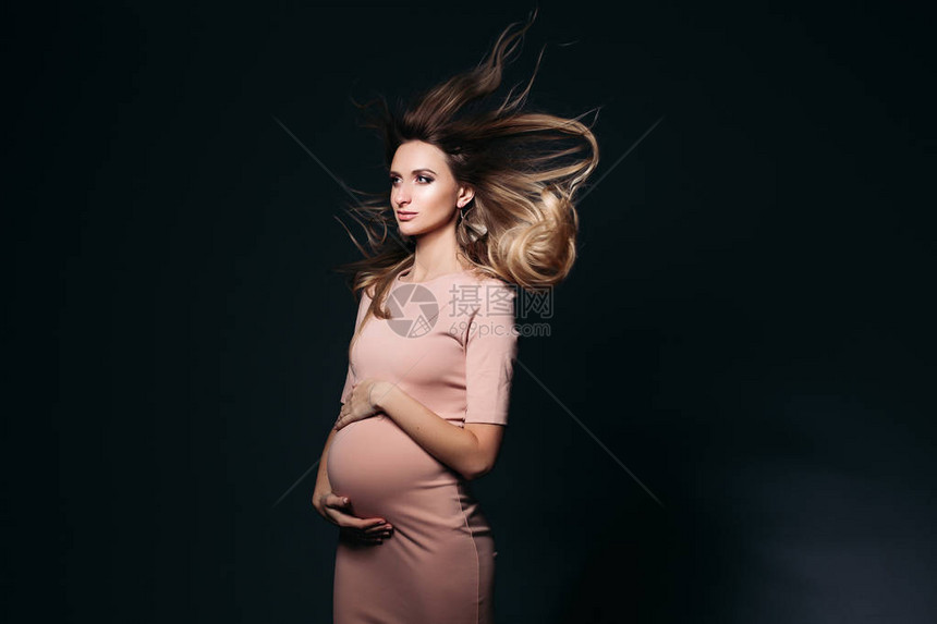 风对穿着粉红色连衣裙拥抱腹部的美丽孕妇的工作室肖像的影响图片