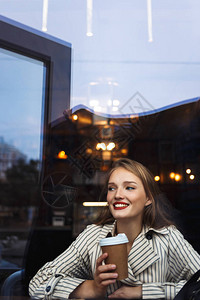 穿着条纹战壕大衣的年轻美丽微笑的女士拿着杯子一起欢乐地在咖图片