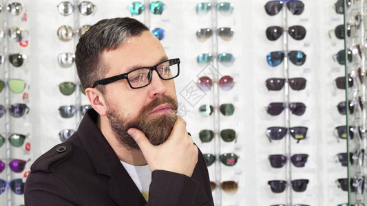 一个成熟的留着胡子的男顾客在眼镜店的显示屏上看着待售的眼镜图片
