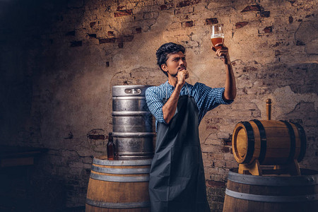 穿着围裙的沉思专家酿酒师站在桶附近检查印地啤酒厂酿图片