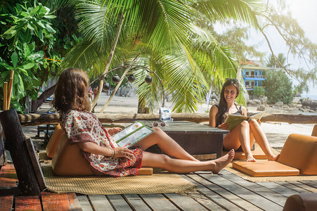 两个享受热带度假的女朋友坐在竹沙滩酒吧咖啡厅看菜单图片