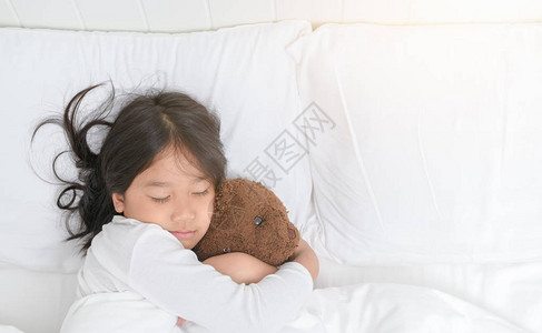 漂亮的小女孩正拥抱着泰迪熊睡在家中的床上高视线是健图片