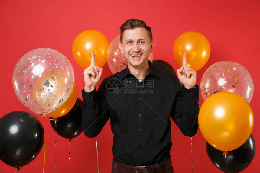 穿着黑色经典衬衫的微笑的年轻人用食指向上指着鲜红色的气球圣瓦伦丁国际妇女节新年快乐生日模拟图片