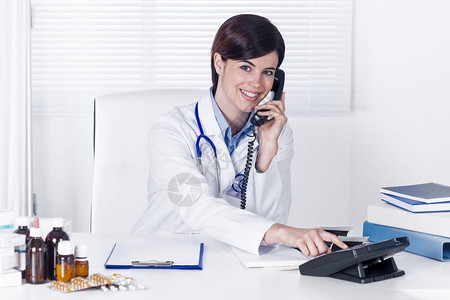 微笑的年轻女医生坐在她的办公桌前拿着药物在她办公室图片