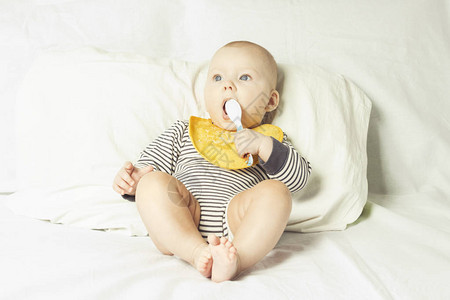 穿着黄色儿童围裙食物残渣手里拿着勺子的小婴儿吃完饭后坐在白色的床上开始喂养婴儿固图片