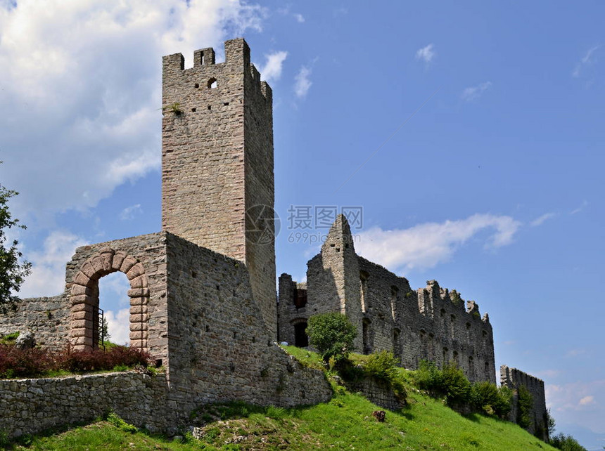 在一座石城堡的入口门上查看带塔的入口门塔由一堵石墙与许多窗户相连城堡位于绿草如茵的山丘上有一些云彩的蓝色天空是城堡上面在Spor图片