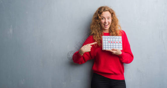 灰色格朗基墙上的年轻红发女人拿着周期日历非常高兴地用图片