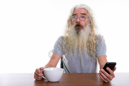 工作室中留胡子老人在用手机和咖啡杯放在木桌上图片