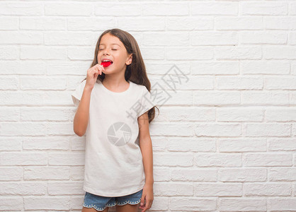 长在白砖墙边的年轻西班牙小孩吃红心棒糖在聪明的面孔图片
