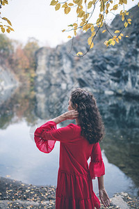 美丽的卷发女郎穿着红礼服看着湖边秋天背景浪图片