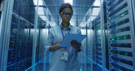 非洲裔美国成年女在数据中心走廊的服务器机架间行走并进行诊断时图片