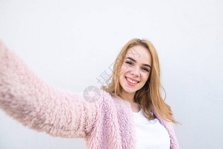 身穿粉色外套和白衬衫的微笑金发女郎站在白墙的背景中图片