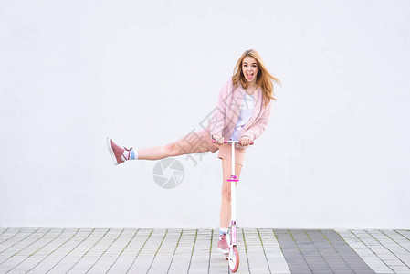 一个穿着粉红色衣服的风趣女孩在白墙的背景上骑着一辆粉红色的滑板车图片