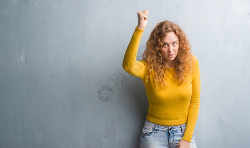 年轻的红发女人在灰色的垃圾墙上愤怒和疯狂地举起拳头沮丧和愤怒图片