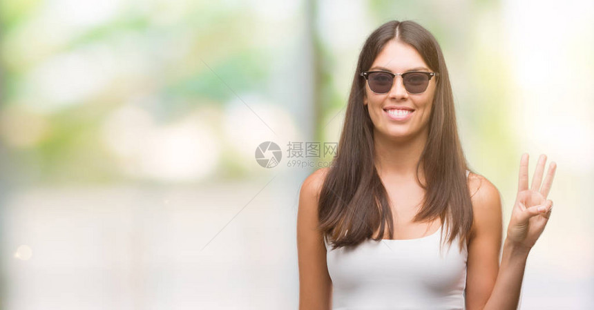 带着太阳眼镜的年轻美丽的西班牙人举起三指头满怀自信和快乐地微笑着容图片