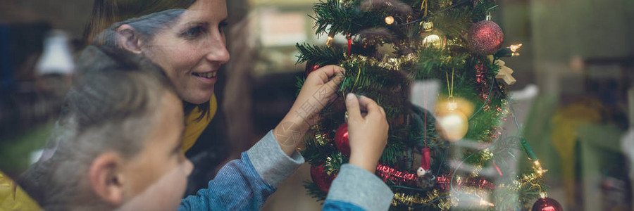 母亲和儿子一起装饰圣诞树的宽视图像通过窗口查看并产生反图片