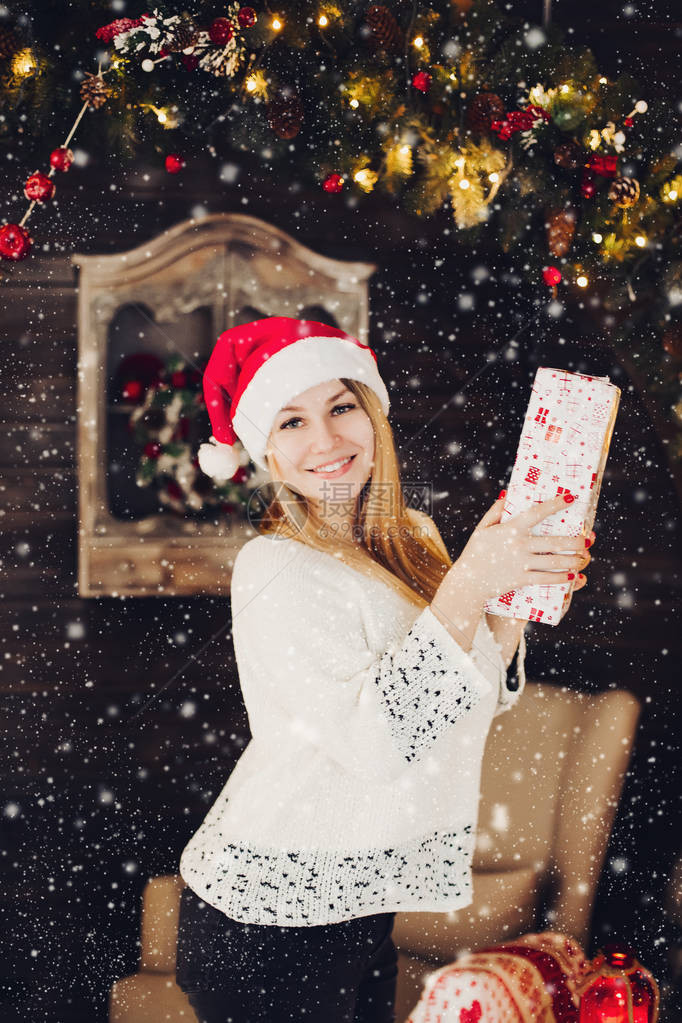 快乐的年轻女子戴着红色的圣诞帽站在装饰着落雪的客厅里图片