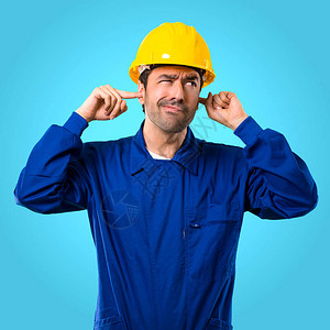年轻工人头戴盔双耳用蓝背景的双手图片