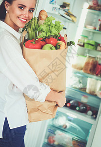 年轻女子拿着杂货购物袋和蔬菜站在厨房里厨房里的图片