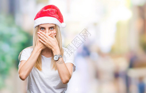 美丽的年轻金发女子的圣诞帽在孤立的上被吓得满嘴胡言乱语地遮住嘴图片
