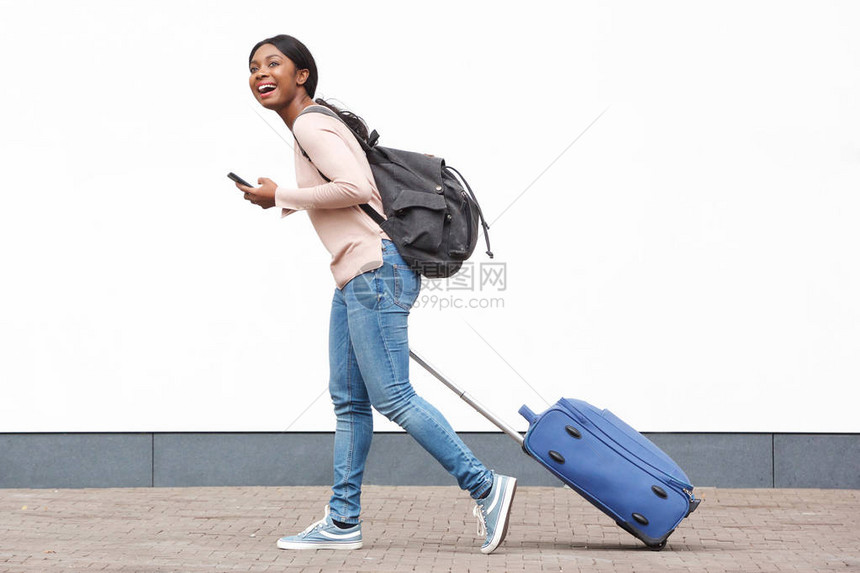 年轻女旅行者用手机和手提箱在白墙上行走的图片