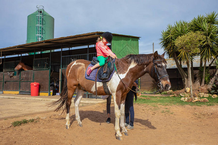 戴着马术帽的四岁金发女孩在一所骑术学校爬一匹白图片