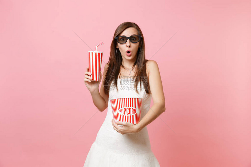 穿着婚纱3d眼镜看电影拿着一桶爆米花塑料杯苏打水或可乐在粉红色柔和的背景上隔离的惊艳新娘婚礼庆典图片