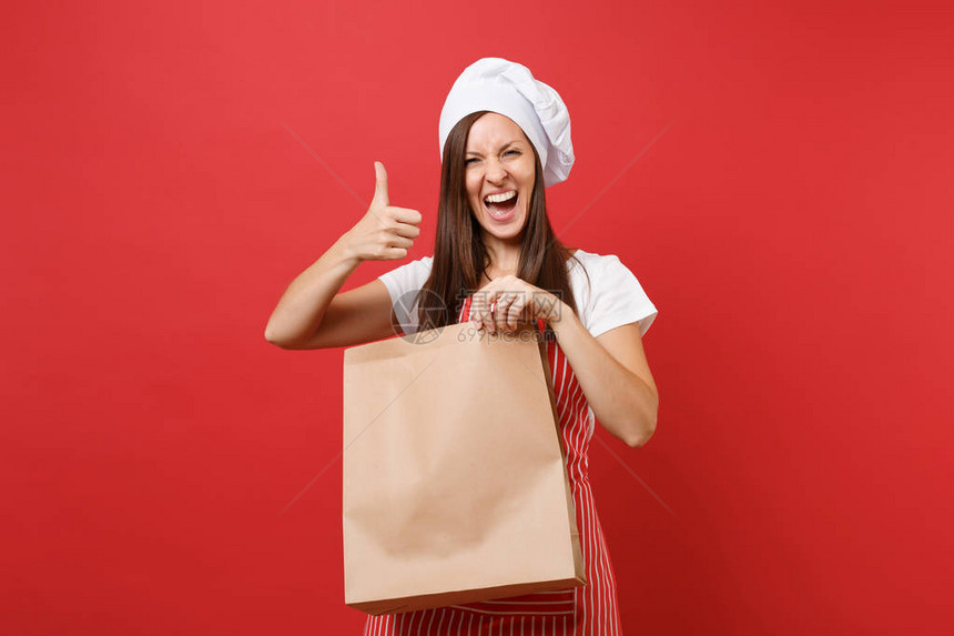 家庭主妇女厨师贝克在条纹围裙t恤无边帽厨师帽隔离在红墙背景女人手握棕色透明空白工艺纸袋外卖模拟图片
