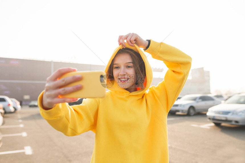 身穿黄色连帽衫的微笑女孩在黄色外壳上自拍智能手机一个穿着街头服装的时尚女人的户图片