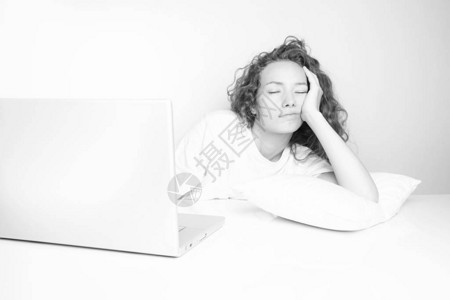 在白墙前用笔记本电脑工作时睡在工作场所的精疲力尽的卷发妇图片