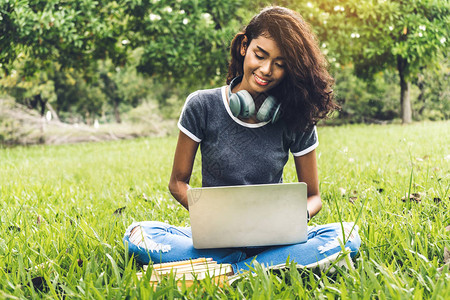 微笑的国际女学生或青少年坐在大学公园里用笔记本电脑做作图片