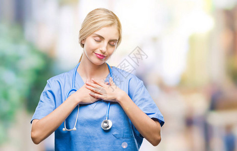 年轻漂亮的金发外科医生护士女人在孤立的背景下微笑着双手放在胸前图片