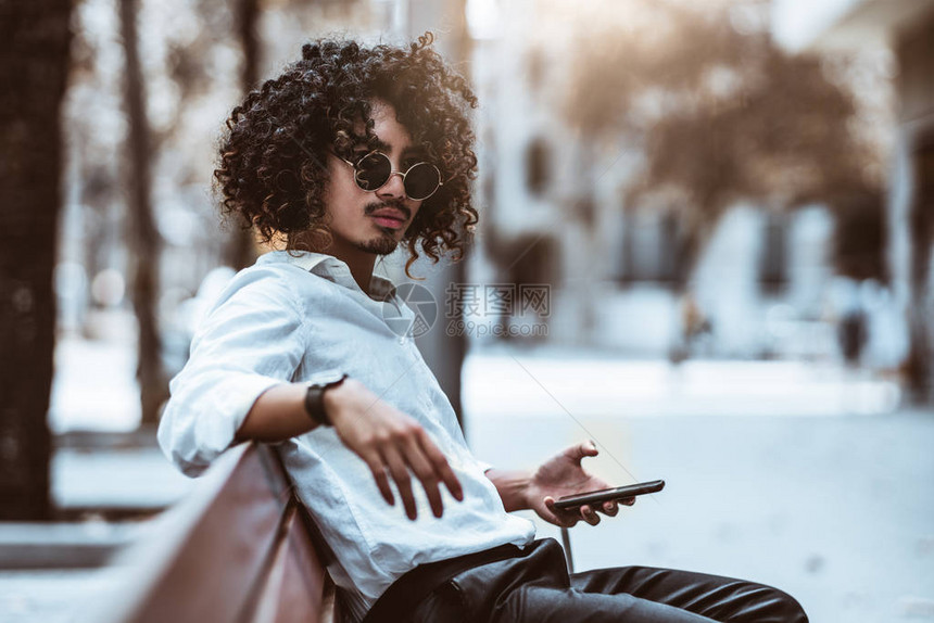 一位戴着墨镜一头卷发坐在公园长椅上手里拿着手机的帅气卷发亚洲男企业家的肖像图片