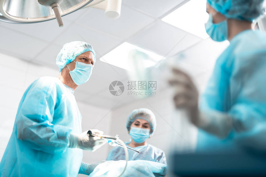 手术室身着无菌袍的中年医生手持腹腔镜仪器而助手站在模糊图片