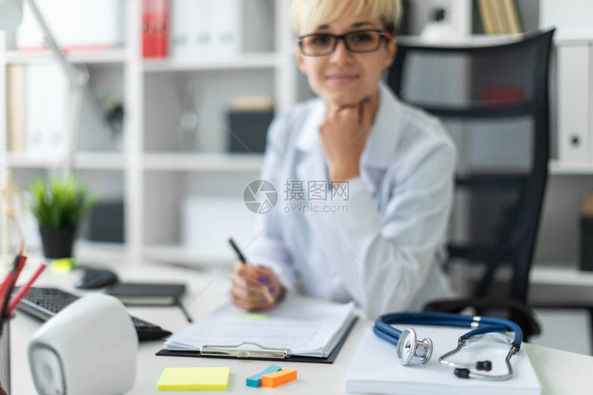 桌子上是一个电话内窥镜一个穿着白色长袍的漂亮年轻女孩在电脑桌的明亮办公室工作她拿着一支笔和一份文件这个女孩留着短白图片