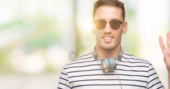 英俊的年轻人戴着耳机展示用第四指头着四号手指笑着自信图片
