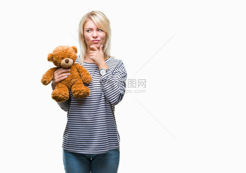 年轻漂亮的金发女郎拿着泰迪熊毛绒在与世隔绝的背景下严肃地思考问题图片