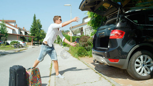 微笑的年轻男游客把一个大手提箱扔进大黑车的后备箱开朗的白人男子从空荡的街道和停在他家门前的汽车后背景图片
