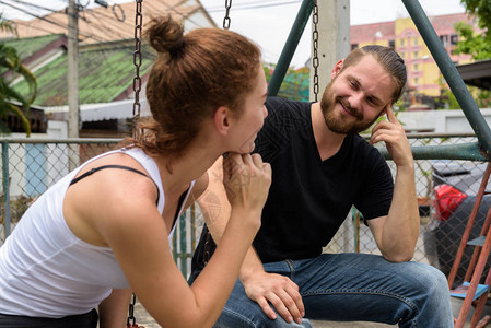 年轻快乐的留着胡子的男人看着年轻漂亮的女人微笑着坐在金属秋千上图片