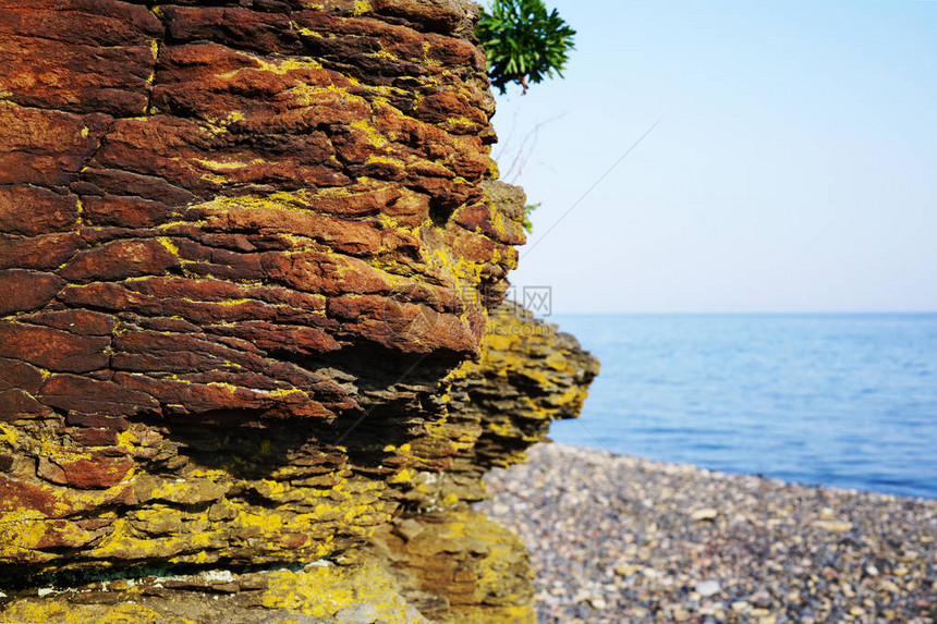 以大海和卵石滩为背景的红褐色岩石特写悬崖后面是海边和碧蓝的海水俄罗斯东部图片