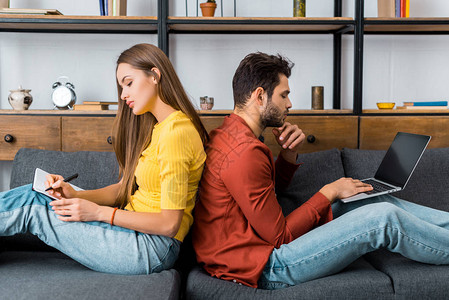 年轻夫妇背靠坐在沙发上用笔记本电脑图片