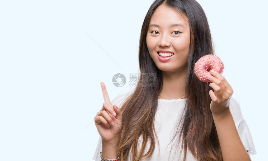 在孤立的背景中吃甜圈的年轻女惊讶于一个想法或问题与快乐的面孔对图片