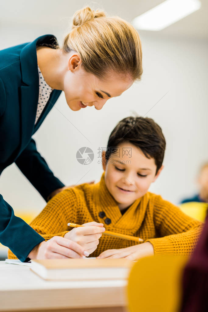 微笑的年轻老师看着小学生用铅笔写字图片