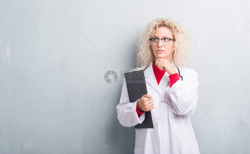 年轻金发女医生在泥黑灰墙上抱着严肃的剪贴板脸认真思考问题非图片