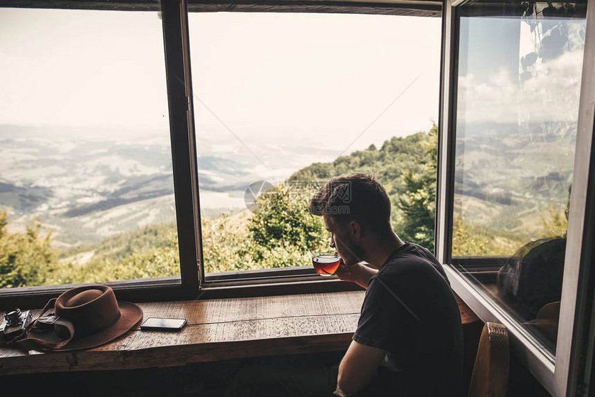 时尚的时髦旅行者在美丽的树林和山脉风景的背景下用玻璃杯喝热茶查看从山上的木窗文本图片