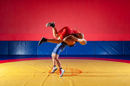 两名身穿红色和蓝色制服的希腊罗马摔跤手在健身房的黄色摔跤地毯上摔跤公图片