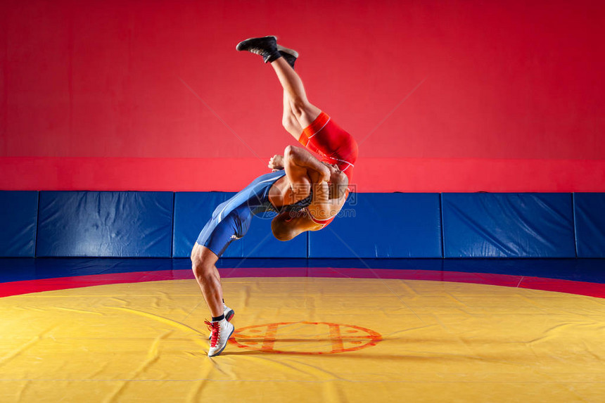 两名身穿蓝色和红色摔跤紧身衣的强壮摔跤手正在健身房的黄色摔跤地毯上摔跤和摔跤做图片