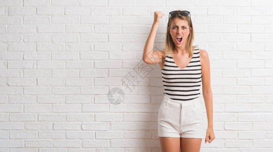 年轻美丽的金发女人在白砖墙上愤怒和疯狂地举起拳头图片
